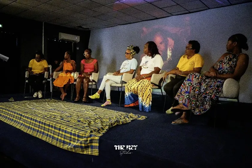Sept personnes assises sur une scène, un tissu madras jaune et bleu étendu devant elles