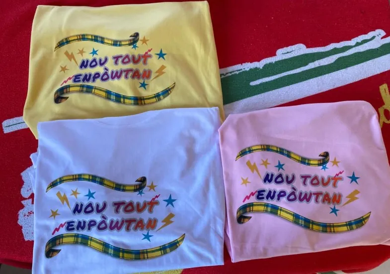 Trois tee-shirts jaune, mauve et rose imprimés « Nou tout’ enpòwtan » et d’un ruban de madras